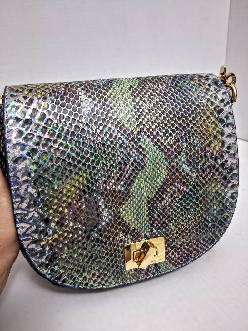 May include: A saddle-shaped shoulder bag featuring a textured snakeskin pattern in iridescent shades of green, blue, purple, and silver. The bag includes a gold-toned metal turn-lock closure on the front flap and gold-toned hardware attachments.