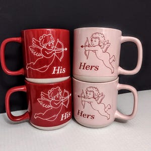 May include: Four ceramic mugs, two red and two pink, stacked in pairs. Each mug features a white illustration of Cupid with a bow and arrow. The red mugs say "His," and the pink mugs say "Hers."