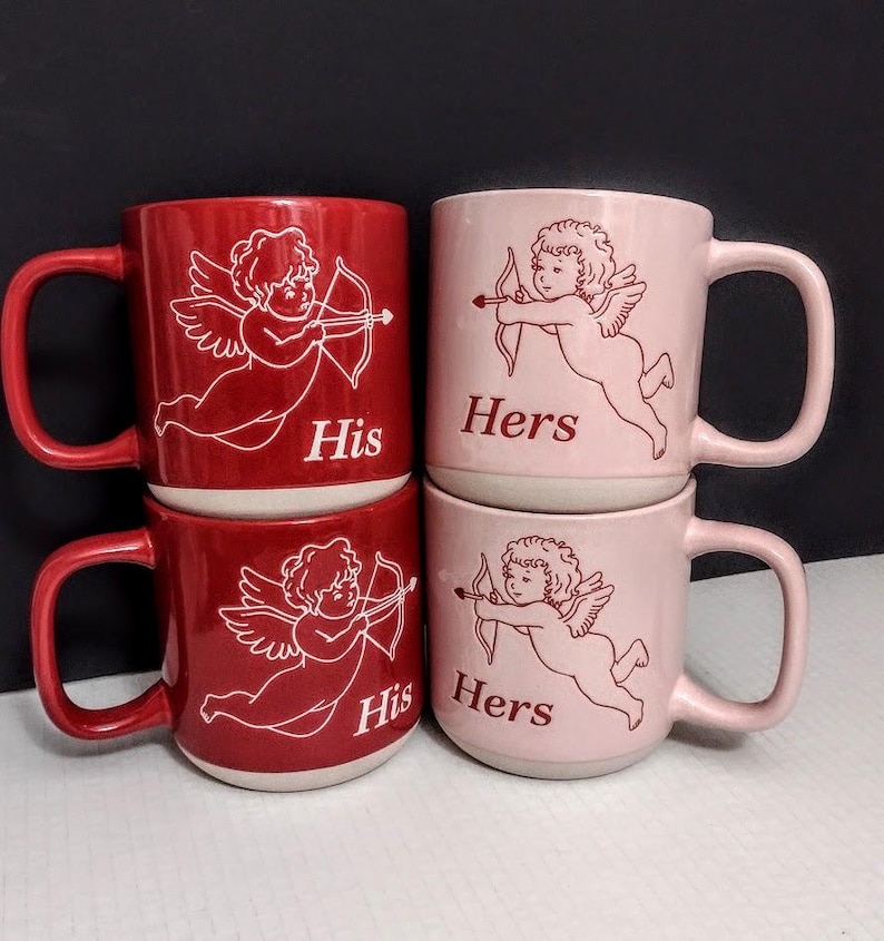 May include: Four ceramic mugs, two red and two pink, stacked in pairs. Each mug features a white illustration of Cupid with a bow and arrow. The red mugs say "His," and the pink mugs say "Hers."