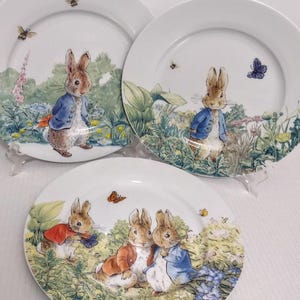 May include: Three white ceramic plates featuring illustrations of Peter Rabbit and other rabbits in a garden setting. The plates have a rim and are decorated with butterflies, flowers, and greenery. The rabbits are wearing jackets.