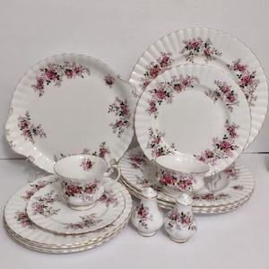 May include: A white porcelain tea set with a pink rose pattern. The set includes plates, saucers, teacups, and salt and pepper shakers. The edges of the plates and saucers have a scalloped design with gold trim.