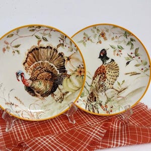 May include: Two decorative plates with a turkey and pheasant design. The plates have a cream-colored background with a yellow rim and are decorated with fall foliage and pumpkins. The plates are displayed on a red and white plaid cloth.