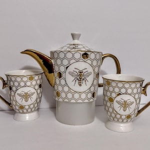 May include: A white and gold bee-themed tea set featuring a teapot and two matching mugs. The teapot and mugs have a honeycomb pattern and a gold bee illustration. The teapot has a gold handle and spout, and the mugs have gold handles.