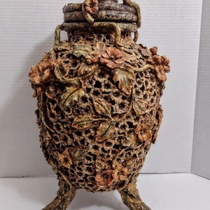 May include: Ornate, brown decorative urn with a lid and three feet. The urn features a floral and leaf design with a lattice-like pattern. The lid has a handle and is also decorated with flowers.