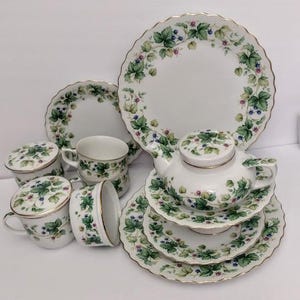 May include: A white porcelain tea set featuring a delicate blackberry and leaf pattern with gold-trimmed scalloped edges. The set includes a teapot with a lid, several cups with matching lids, and multiple plates of varying sizes, all decorated consistently.