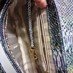 May include: A handbag with an iridescent snakeskin pattern in shades of green, blue, and purple. The interior features a beige and brown striped lining, a zipped pocket, and a gold-toned metal lobster clasp attached to a matching leather strap.