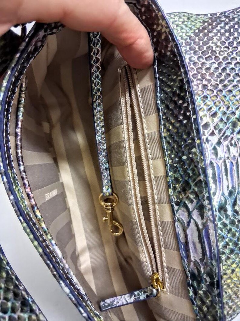 May include: A handbag with an iridescent snakeskin pattern in shades of green, blue, and purple. The interior features a beige and brown striped lining, a zipped pocket, and a gold-toned metal lobster clasp attached to a matching leather strap.