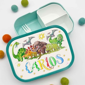 Könnte beinhalten: Eine türkis-weiße Lunchbox mit Dinosaurier-Motiv und dem Namen "CARLOS" in bunten Buchstaben. Der Deckel zeigt eine Cartoon-Illustration verschiedener Dinosaurier, eine Sonne und Sterne. Enthält eine weiße Gabel und einen geteilten Behälter.