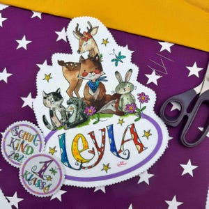 Könnte beinhalten: Eine skurrile Illustration mit Waldtieren, darunter ein Reh, ein Fuchs und ein Hase, mit dem Namen "Leyla" in bunten Buchstaben. Zwei runde Abzeichen mit der Aufschrift "Schul Kind 2022" und "1 Klasse". Der Hintergrund ist lila mit weißen Sternen.
