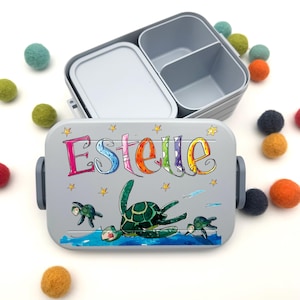Könnte beinhalten: Eine graue Lunchbox mit abnehmbarem Deckel. Die Lunchbox hat ein Fach für ein Sandwich und ein kleineres Fach für Snacks. Die Lunchbox ist mit einer bunten Illustration von Meeresschildkröten und dem Namen "Estelle" in Regenbogenfarben verziert.