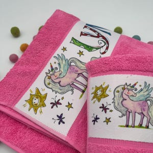 Könnte beinhalten: Zwei rosa Handtücher mit Einhorn-Motiv. Ein Handtuch zeigt den großen Buchstaben "M" in bunten Buchstaben. Beide Handtücher haben einen weißen Rand mit einem Einhorn-, Sonnen- und Stern-Design. Die Handtücher sind aus weichem, saugfähigem Material.