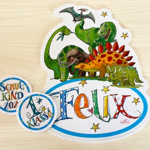 Op de afbeelding: Een witte vilt-ovaal met een blauwe rand en de naam "Felix" geschreven in kleurrijke letters. De ovaal is versierd met sterren en omringd door een kleurrijke illustratie van dinosaurussen, waaronder een groene Tyrannosaurus Rex, een bruine Stegosaurus, een groene Triceratops en een bruine Brachiosaurus. Er zijn twee kleinere cirkels met blauwe randen en de tekst "Schul Kind 202" en "1. Klasse" geschreven in kleurrijke letters.