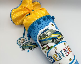 May include: A blue and yellow school cone with a police car design and the name "Benn" embroidered on the front. The cone has blue pom-poms and white polka dots. It also has a small tag with the text "Schul Kind 20" on it.