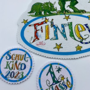 Könnte beinhalten: Ein Set aus drei Filzpatches mit Dinosaurier-Motiv. Der größte Patch zeigt einen grünen Tyrannosaurus Rex und einen grünen Triceratops mit dem Wort "Finer" in bunten Buchstaben. Die beiden anderen Patches sind rund und zeigen die Wörter "Schulkind 2023" und "1. Klasse" in bunten Buchstaben.