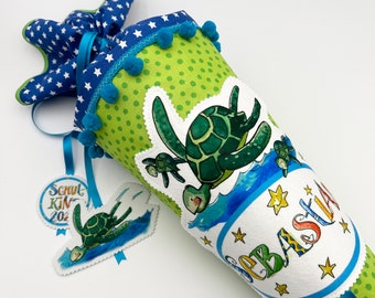 May include: A green and blue school cone with a turtle theme. The cone has a white fabric panel with the name "Sebastian" written in colorful letters. The cone is decorated with blue pom-poms and stars. A small tag with a turtle design and the text "Schul-Kin 202" is attached to the cone.