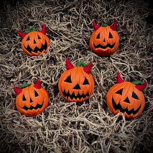 May include: Five orange pumpkin decorations with devil horns and carved faces. The pumpkins have black eyes and mouths, red horns, and green stems. They are arranged on a bed of dried, light brown plant material.