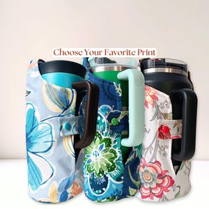 Può includere: Tre bicchieri termici con manici, ciascuno rivestito in una custodia di tessuto con distinti motivi floreali nei toni del blu, verde e crema. Il testo "Choose Your Favorite Print" è visualizzato sopra gli articoli, dotati di bottoni a pressione.