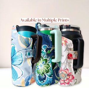 May include: Three insulated tumblers with handles, each encased in a fabric sleeve featuring distinct floral patterns. The text Available in Multiple Prints is centered above the tumblers. The sleeves are secured with small colored snaps.