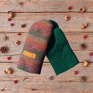 May include: A pair of felted wool mittens featuring one mitten with a variegated pattern of orange, green, and purple tones and one solid dark green mitten. A small rectangular tan leather tag with text is attached to the patterned mitten.