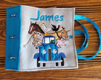 May include: A blue felt book with a blue felt strap. The book has a gray felt cover with a blue truck, a frog, and various farm animals embroidered on it. The book is personalized with the name "James".
