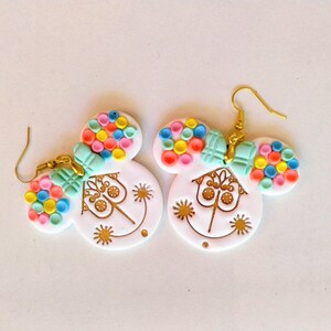May include: A pair of white, whimsical earrings with a Mickey Mouse-inspired design. Each earring features a round face with a gold-colored smiling face and a colorful, round cluster of beads. The earrings have gold-colored hooks and a light blue bow.