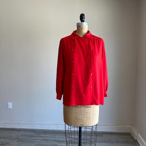 May include: A bright red long-sleeved blouse with a button-up front and a rounded collar. The blouse is displayed on a vintage dress form with a wire cage base. The fabric appears to be a smooth, lightweight material.
