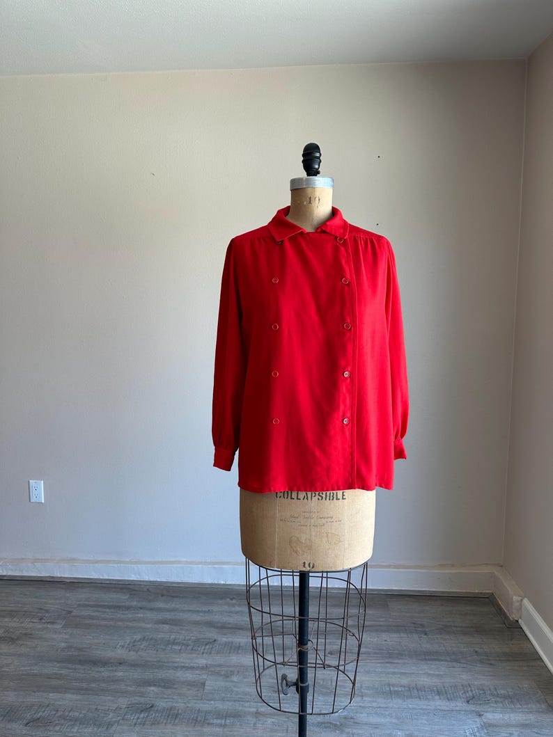 May include: A bright red long-sleeved blouse with a button-up front and a rounded collar. The blouse is displayed on a vintage dress form with a wire cage base. The fabric appears to be a smooth, lightweight material.