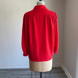May include: A vibrant red blouse with long sleeves and a classic collar, displayed on a vintage dress form. The back view showcases the blouse's tailored design and buttoned cuffs, perfect for a vintage clothing collection.