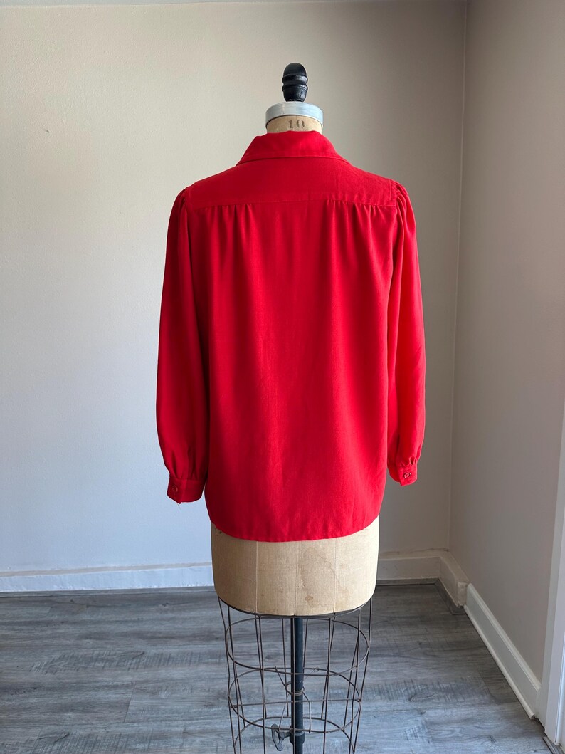 May include: A vibrant red blouse with long sleeves and a classic collar, displayed on a vintage dress form. The back view showcases the blouse's tailored design and buttoned cuffs, perfect for a vintage clothing collection.