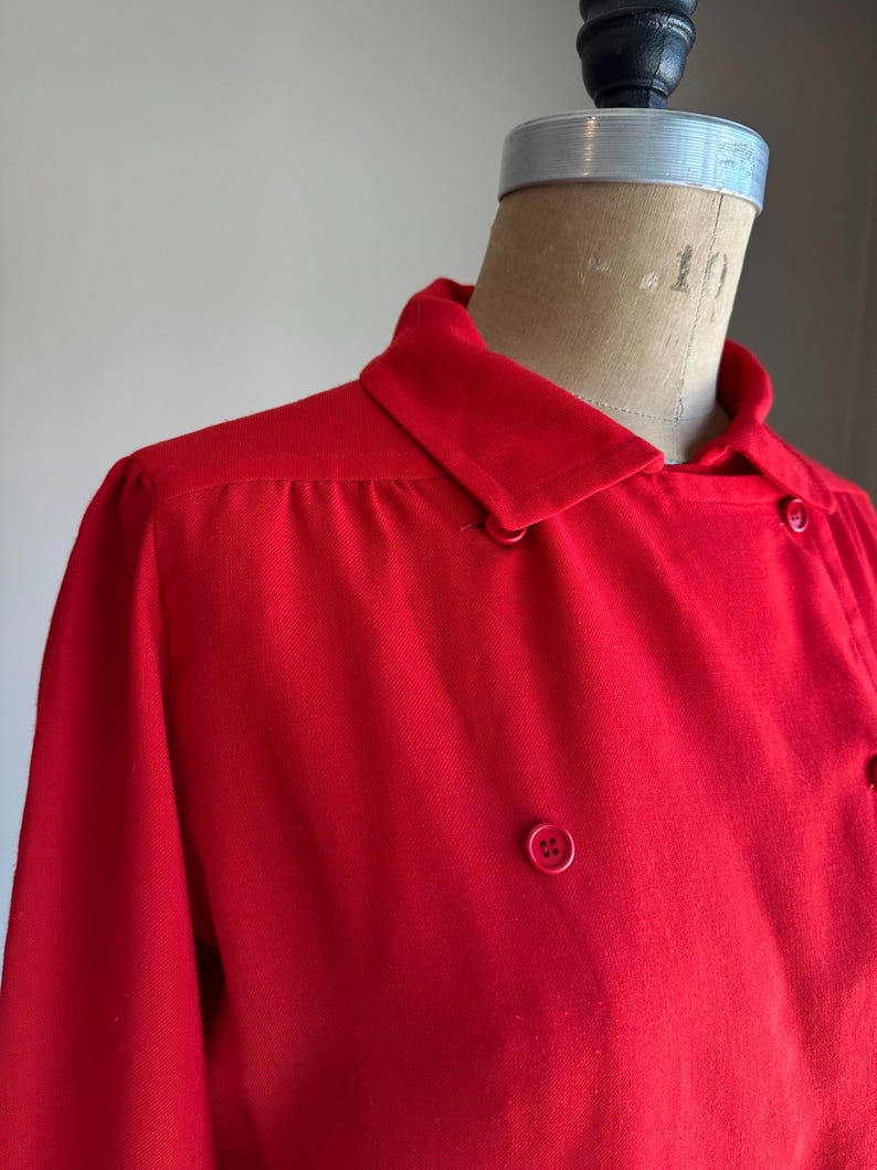 May include: A bright red collared jacket with a double-breasted design. The jacket features a classic collar, long sleeves, and several matching red buttons. The garment is displayed on a mannequin.