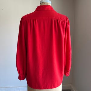 May include: A bright red long-sleeved blouse with a classic collar and buttoned cuffs. The back view of the blouse is displayed on a mannequin. The fabric appears to be a smooth, solid color.
