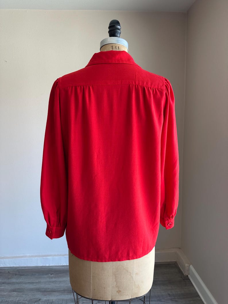 May include: A bright red long-sleeved blouse with a classic collar and buttoned cuffs. The back view of the blouse is displayed on a mannequin. The fabric appears to be a smooth, solid color.