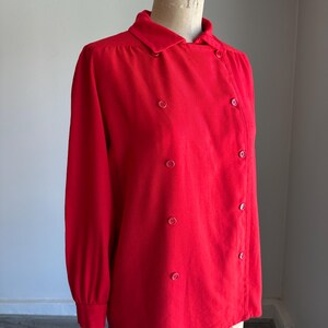 May include: A bright red long-sleeved blouse with a double-breasted design. The blouse features a collared neckline and is fastened with round, matching red buttons. The garment is displayed on a mannequin.
