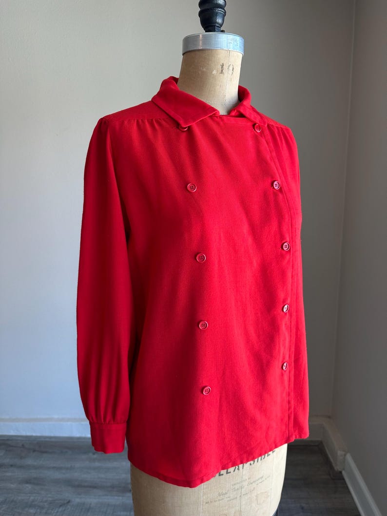 May include: A bright red long-sleeved blouse with a double-breasted design. The blouse features a collared neckline and is fastened with round, matching red buttons. The garment is displayed on a mannequin.