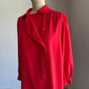May include: A bright red double-breasted blouse with long sleeves and a collared neckline. The blouse is buttoned up and displayed on a mannequin. The fabric appears to be a smooth, solid color.
