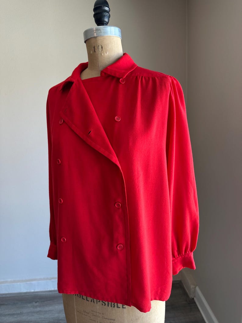 May include: A bright red double-breasted blouse with long sleeves and a collared neckline. The blouse is buttoned up and displayed on a mannequin. The fabric appears to be a smooth, solid color.