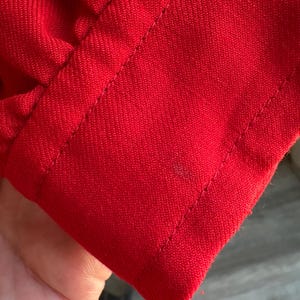 May include: Close-up of a vibrant red fabric, possibly a sleeve or cuff, with visible stitching along the edges. The fabric has a textured appearance, and the stitching is neatly done, highlighting the garment's construction.