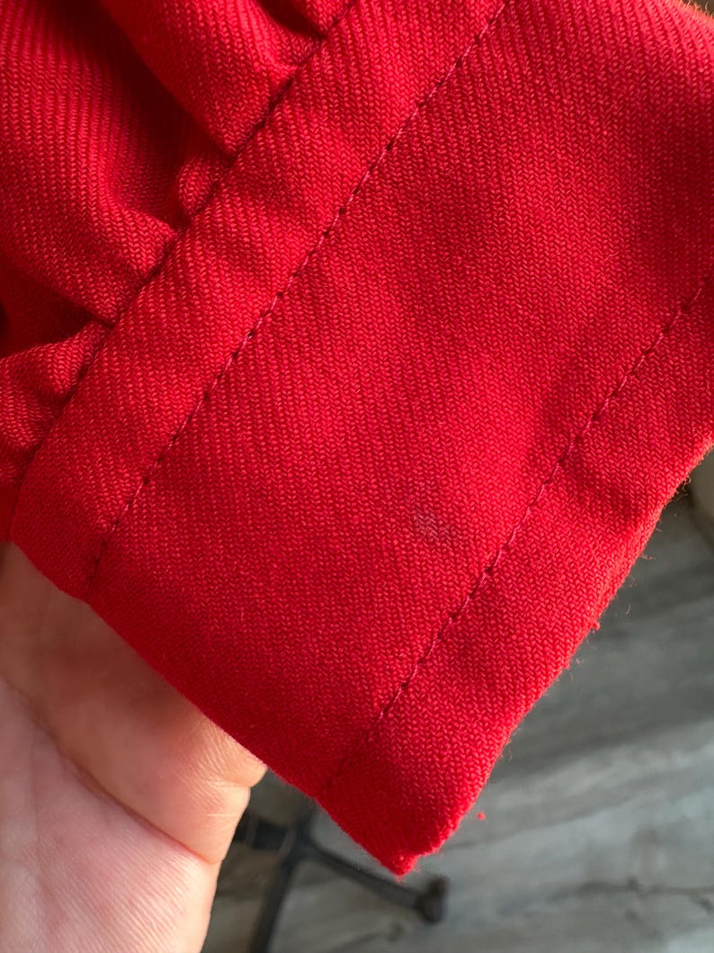 May include: Close-up of a vibrant red fabric, possibly a sleeve or cuff, with visible stitching along the edges. The fabric has a textured appearance, and the stitching is neatly done, highlighting the garment's construction.