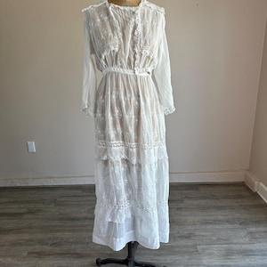 May include: A vintage white dress with long sleeves and intricate lace detailing. The dress features a high neckline, a fitted waist, and a tiered skirt. It is displayed on a vintage dress form, showcasing its delicate design and historical style.