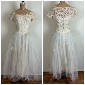 May include: A vintage white lace wedding dress with a tulle skirt and a fitted bodice. The dress has a scoop neckline and short sleeves. The bodice is decorated with delicate lace.