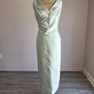 May include: A pale green, floor-length dress with a V-neckline. The dress is displayed on a vintage mannequin. The neckline is embellished with silver-toned beading. The dress has a simple, elegant design.
