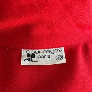 May include: Close-up of a bright red garment with a textured weave. A white label with the text "courr&egrave;ges paris" and a logo is visible. The garment appears to be a piece of clothing.