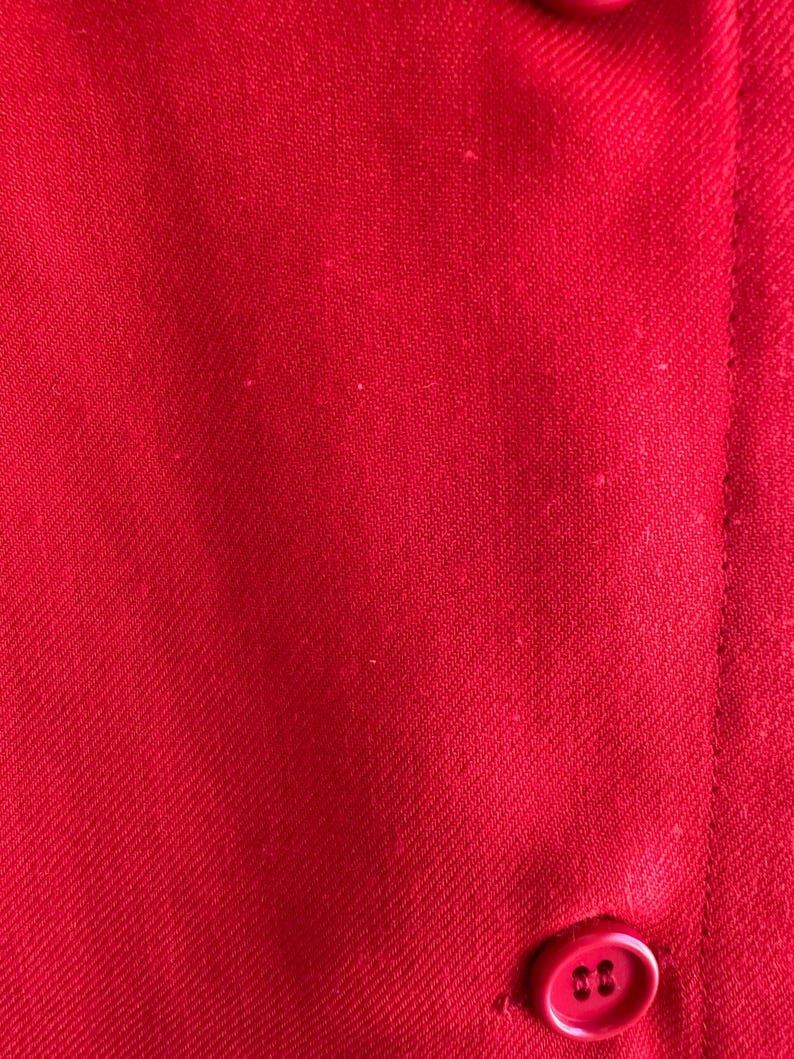 May include: Close-up of a vibrant red fabric with a subtle woven texture. The image shows a portion of a garment, with a button and a seam visible. The fabric's color is consistent throughout the image.