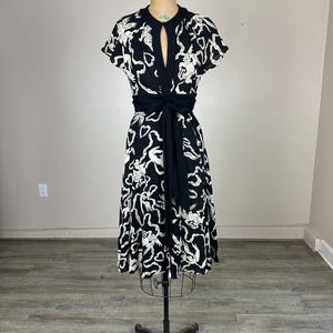 May include: Black and white patterned dress with a keyhole neckline and short sleeves. The dress features a black waist tie and is displayed on a vintage dress form. The pattern appears to be a repeating design of abstract shapes.