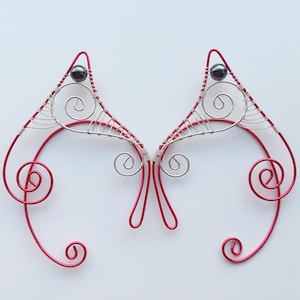 May include: Two red wire elf ear cuffs, each with a triangular top and a dark bead. The design incorporates silver wire detailing and decorative swirls. These fantasy-themed ear cuffs are a unique accessory.