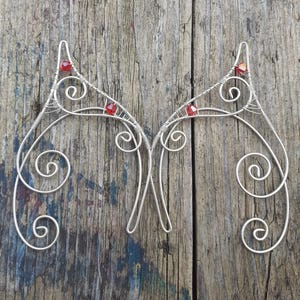 May include: Two silver wire ear cuffs with red gemstone details. The ear cuffs have a delicate, swirling design and are displayed on a weathered wooden surface. The design is reminiscent of elven ears.