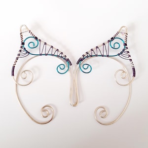 May include: A pair of silver-coloured wire elf ear cuffs with teal and purple accents. The ear cuffs feature a delicate, handcrafted design with swirling details and a pointed ear shape. The wire is intricately woven, creating a unique and whimsical accessory.