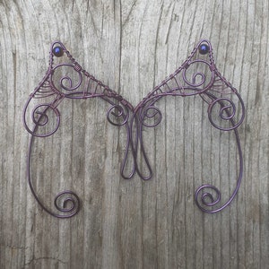 May include: A pair of purple wire fairy wings with intricate scroll and swirl designs. Each wing features a small, dark blue bead at the top. The wings are displayed against a weathered wooden background.