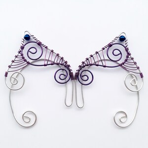 May include: A pair of handcrafted wire ear cuffs. The ear cuffs have a silver base with purple wire accents and small blue beads. The design includes swirls and geometric shapes, creating an elegant, fantasy-inspired accessory.