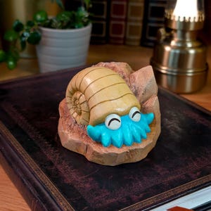 May include: A carved figurine of an ammonite fossil featuring a tan, spiraled shell and a bright turquoise creature with closed eyes emerging from the front, all set upon a textured, rock-like base with a light brown finish and carved details.